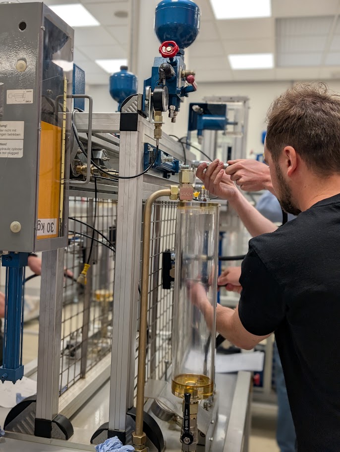 Zwei Personen arbeiten in einem Technikraum an einer industriellen Versuchsanlage mit transparentem Zylinder, Ventilen, Rohren und Messgeräten.