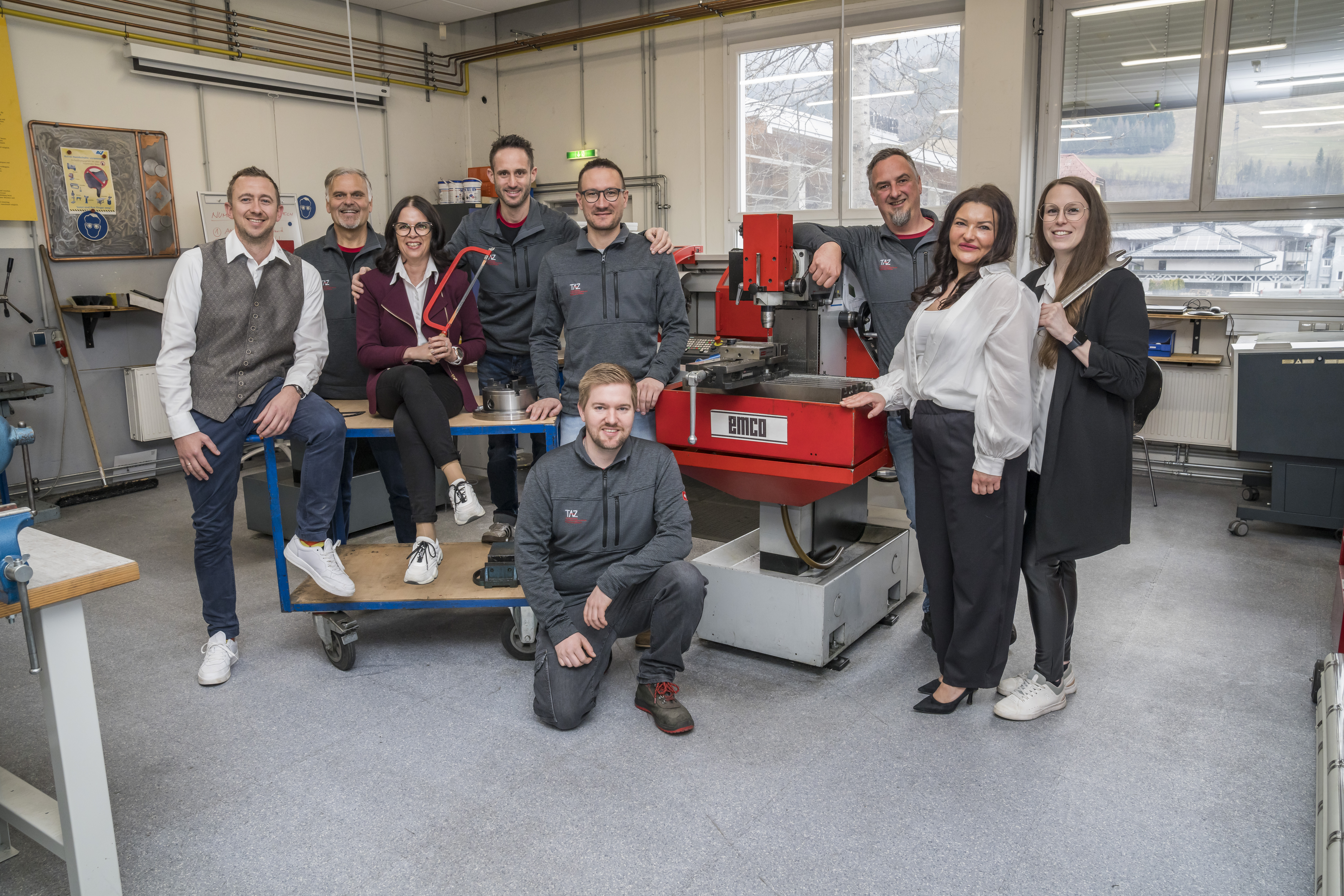 In einer Werkstatt stehen mehrere Menschen (Männer und Frauen) um eine Maschine. Sie tragen zum Teil Arbeitskleidung und scheinen in einem technischen Umfeld zu arbeiten.